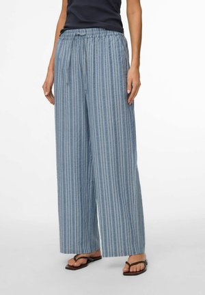 Inimene, kes kannab siniste ja valgete vertikaalsete triipudega laia säärega pükse, millel on nöörvöökoht, ning musti plätusid, seisab valgel taustal.