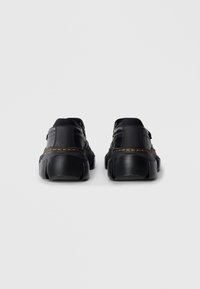 O pereche de pantofi negri din piele cu talpă groasă și cusături galbene, văzuți din spate pe un fundal simplu deschis.