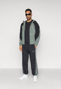 Športový outfit s čierno-zelenou bundou so zipsovými vreckami a zodpovedajúcimi čiernymi nohavicami so zelenými bočnými akcentmi. Biely tenisky a slnečné okuliare.