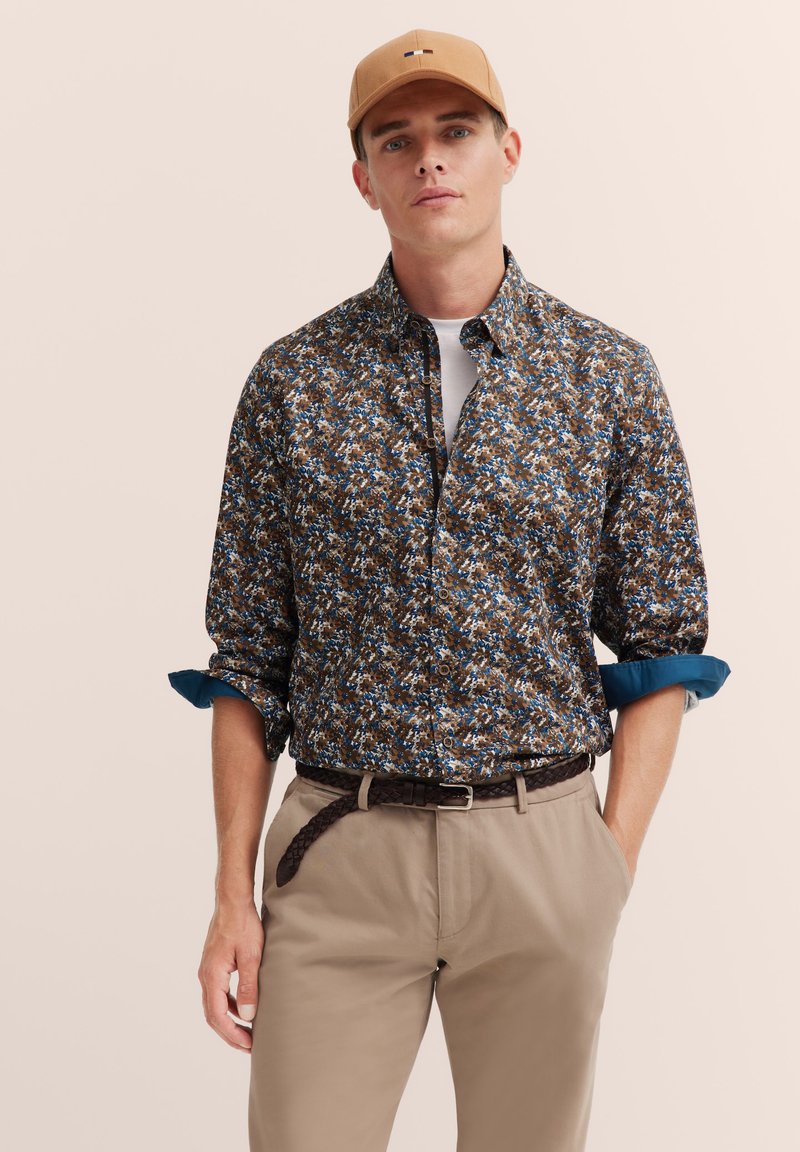 Braun geblümtes Hemd mit Knöpfen und blauen Akzenten, kombiniert mit beigen Hosen und einem braunen geflochtenen Gürtel. Eine lässige Mütze rundet den Look ab.