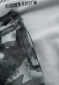 Tessuto grigio con motivo mimetico in bianco e nero. Presenta una cintura elastica e un dettaglio di tasca laterale, mostrando una combinazione di trame.