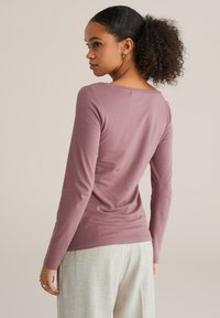 Ilgstoša piedurkne augšdaļa muted mauve audumā ar apaļu izgriezumu un pieguļošu siluetu, kopā ar gaiši pelēkiem, teksturētiem biksēm.