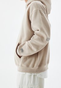 Haut à capuche beige en tissu doux, avec une poche kangourou, des poignets côtelés et un patch logo sur la manche. Texture lisse.