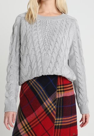 Pull en maille torsadée gris clair avec un décolleté large et un motif texturé, associé à une jupe à carreaux présentant des teintes rouges, marines et jaunes.