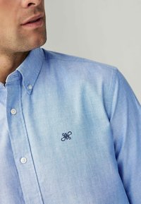 Camisa de botón azul claro con cuello, con botones blancos y un logotipo bordado en azul marino en el pecho; textura de tela suave.