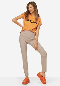 Pruhované tričko v oranžové a žluté barvě s grafikou "vlna"; béžové slim-fit kalhoty; oranžové platformové sandály na klínku. Hladké textury po celém outfitu.