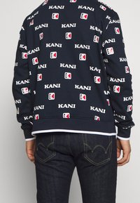 Felpa navy con motivo ripetuto in bianco della scritta "KANI" e del logo. Tessuto morbido con polsini e orlo a coste; abbinata a jeans in denim scuri.