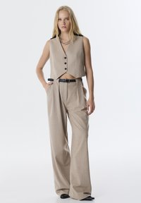Beige cropped vest met zwarte schouderaccenten en vijf knopen, gecombineerd met hoog-getailleerde beige broeken met subtiele stippen en een zwarte riem.
