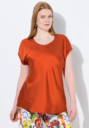 T-shirt en soie couleur rouille avec un col rond et des manches courtes roulées ; texture lisse. Associé à un pantalon à motifs présentant des designs de fruits.