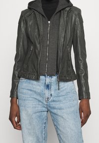 Zwarte leren motorjack met ritssluitingen en geribbelde details, gelaagd over een grijze rits hoodie en gecombineerd met lichtblauwe jeans.