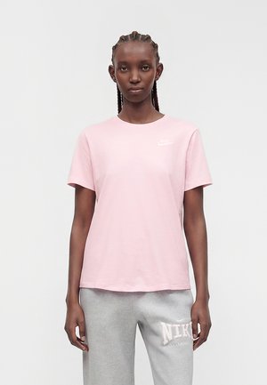 Kvinde med fletninger står iført en lyserød Nike T-shirt og lysegrå Nike joggingbukser mod en ensfarvet hvid baggrund.