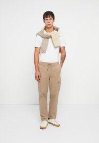 Pantalons cargo beige avec poches latérales, associés à un t-shirt blanc. Un pull en tricot gris-beige est drapé sur les épaules. Baskets blanches.