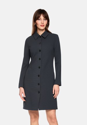 Frau trägt ein langärmliges, knöpfbares, dunkelgraues Kleid mit Kragen und steht vor einem schlichten weißen Hintergrund.