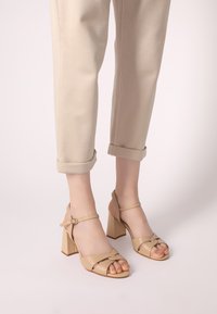 Beige High-Impact Sandalen mit Knöchelriemen, offener Zehenpartie und Blockabsatz. Glatte Textur mit gekreuztem Vorderriemen-Design. Kombiniert mit hochgekrempelten beigen Hosen.