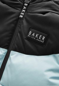 Juodas ir šviesiai mėlynas paminkštintas švarkas, turintis lygų paviršių, užtrauktuką ir logotipo pleistrą su užrašu "BAKER by TED BAKER."
