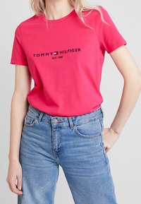 Camiseta de algodón rosa con el logo de "Tommy Hilfiger" en negro, combinada con jeans de denim azul claro. Diseño clásico de cuello redondo.