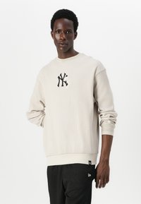 Lys beige ribbesweater med rund hals, med en svart New York-logo på brystet og en svart etikett på nederkanten.