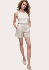 Beige Leinen-Shorts mit maßgeschneidertem Design, hoher Taille und ausgefranstem Saum, kombiniert mit einem taillierten weißen ärmellosen Oberteil und Slip-On-Schuhen.
