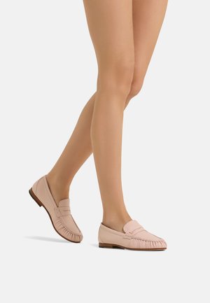 Gambe che indossano mocassini in pelle rosa chiaro con dettagli di cuciture e tacchi bassi in legno, posate come in movimento su sfondo bianco uniforme.