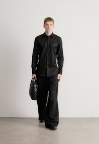 Fekete hosszú ujjú ing és bőszárú nadrág, fekete bőrtáskával párosítva, sima textúrával és testhezálló sziluettel.