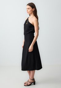 Schwarzes, einseitiges Kleid in figurbetonter Passform mit fließendem Rock. Hergestellt aus einem glatten Stoff, kombiniert mit schwarzen, rückenfreien Sandaletten mit Absatz.