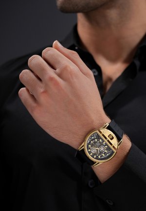 Mann in schwarzem Hemd zeigt sein Handgelenk mit einer goldenen und schwarzen Skelettuhr mit einem einzigartigen, geschwungenen, dreieckigen Gehäuse und schwarzem Armband.