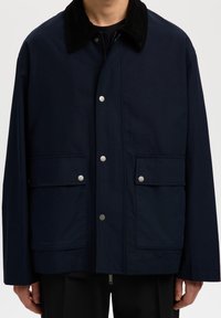 Homme portant une veste bleu marine avec un col en velours côtelé noir, des boutons-pression à l'avant, deux grandes poches à rabat et une fermeture éclair en dessous.