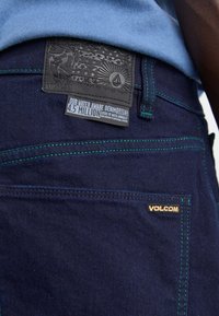 Tamno plavi traperici imaju svijetlozelene šavne, teksturiranu crnu kožnu oznaku s reljefnim detaljima i zlatni logo "VOLCOM".