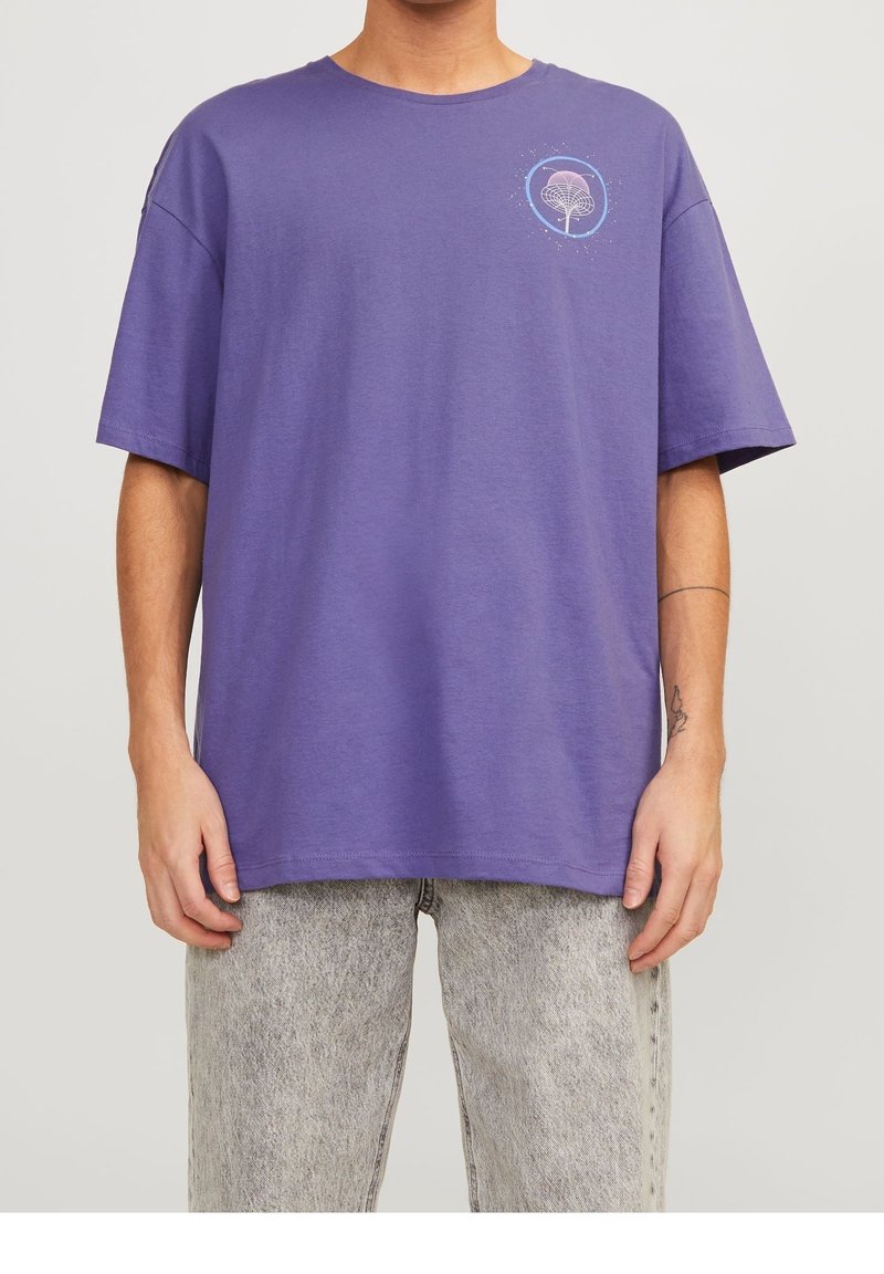 Homme portant un T-shirt violet ample avec un motif blanc abstrait en haut à gauche, associé à un jean gris clair délavé à l'acide et un tatouage sur le bras.