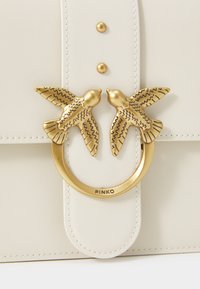 Λευκή δερμάτινη τσάντα με χρυσό κυκλικό κούμπωμα που απεικονίζει δύο λεπτομερείς πουλιά, συμπληρωμένη από χρυσά διακοσμητικά νήματα πάνω από το κούμπωμα.