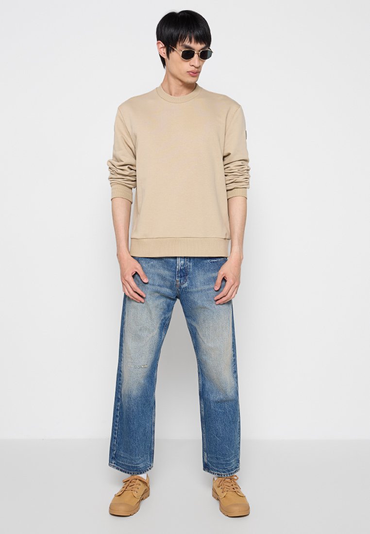 Felpa beige con orlo a coste, maniche morbide; abbinata a jeans ampi in denim blu sbiadito. Sneakers marroni e occhiali da sole scuri completano il look.