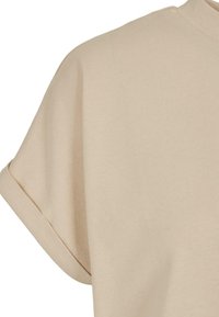 Chemise beige à manches courtes avec poignets retroussés et col rond, montrée depuis l'épaule et la partie supérieure de la poitrine.