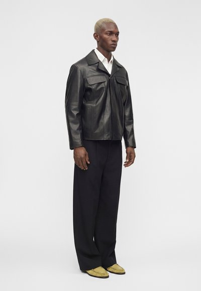 Mannequin masculin aux cheveux blonds courts porte une veste en cuir noire, une chemise blanche, un pantalon noir large et des mocassins jaunes, debout devant un fond uni.