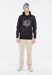 Zwarte hoodie met wit geometrisch ontwerp en logo, gecombineerd met beige broek en grijze beanie. Bevat een trekkoord en ribgebreide manchetten.