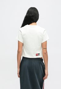 Κοντομάνικη λευκή μπλούζα cropped, με μικρή ετικέτα στην πλάτη που έχει λογότυπο και μαύρες και κόκκινες λεπτομέρειες. Υφή υφάσματος.