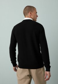 Hombre con cabeza afeitada que lleva un suéter negro texturizado, camisa blanca con cuello y pantalones beige, de espaldas contra un fondo gris plano.