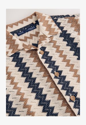 Lyhythihainen kudottu paita, jossa on siksak-kuvio kerman, ruskean ja laivastonsinisen sävyissä. Paidassa on kaulus ja napit yksityiskohtina. Yleisilme pehmeä tekstuuriltaan.