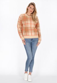 Beige gebreide trui met oranje en bruine ruiten, relaxed fit, ribgebreide manchetten, gecombineerd met lichtblauwe skinny jeans en witte sneakers.