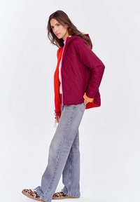 Kobieta ubrana w pikowaną bordową kurtkę z pomarańczowym wykończeniem, jasnoszare dżinsy i trampki w panterkę, stojąca na tle jednolitego białego tła.