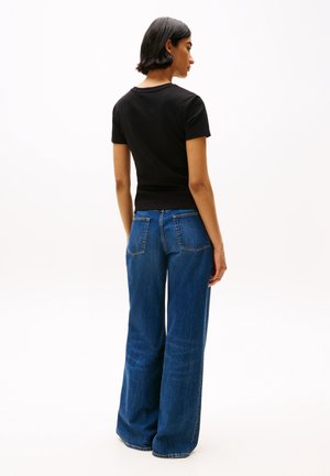 Rövid sötét hajjal rendelkező nő fekete rövid ujjú ingben és kék, bő szárú farmernadrágban, háttal áll egy egyszerű háttér előtt.
