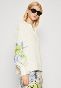 Witte blouse met lange, opgeblazen mouwen. Voorzien van kleurrijke bloemenborduurwerk in blauw en groen op de linker mouw. Soepele textuur.