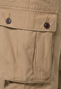 Beige katoenen stof met een groot, met knopen gesloten zakje. Heeft stiksel details en een gestructureerd oppervlak. De knopen zijn donkerbruin met vier gaatjes.