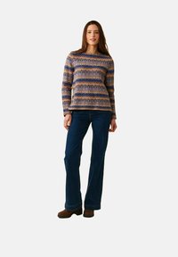 Kvinna som står och bär en långärmad mönstrad tröja, mörkblå jeans och bruna skor mot en enkel ljus bakgrund.