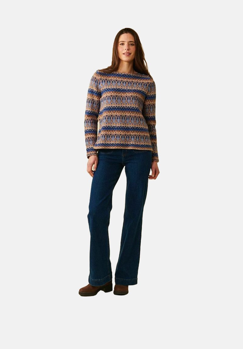 Kvinna som står och bär en långärmad mönstrad tröja, mörkblå jeans och bruna skor mot en enkel ljus bakgrund.