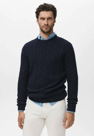 Suéter de punto trenzado en navy con puños y dobladillo de canalé, llevado sobre una camisa de cuello azul claro, combinado con pantalones blancos. Detalle de diseño texturizado visible.