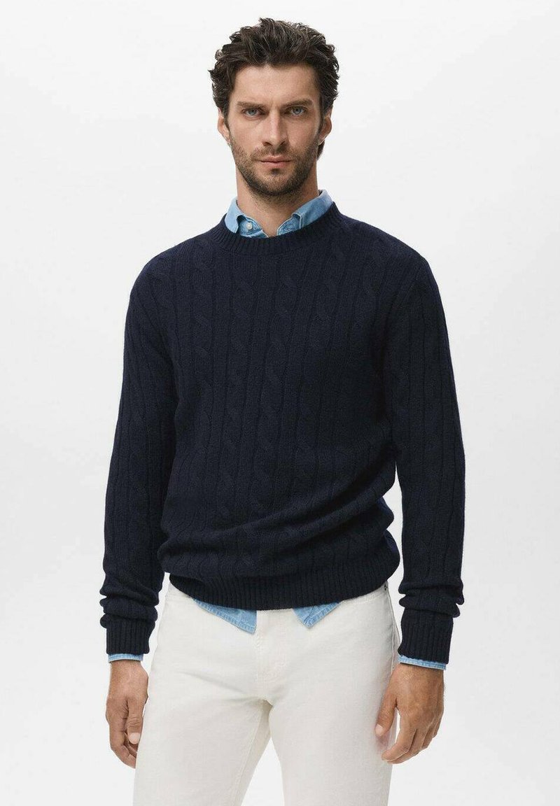 Suéter de punto trenzado en navy con puños y dobladillo de canalé, llevado sobre una camisa de cuello azul claro, combinado con pantalones blancos. Detalle de diseño texturizado visible.