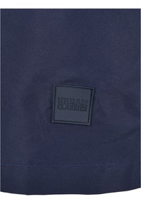 Temno moder tkanina z teksturirano površino; vsebuje kvadratno črno oznako z vtisnjenim logotipom "URBAN CLASSICS" na dnu v sredini.