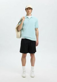 Ljusblå pikétröja, svarta shorts, vita sneakers och en beige keps. Håller en beige toteväska. Enkel, avslappnad stil, mjuk textur.