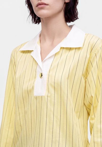 Femme portant une chemise jaune à manches longues avec de fines rayures verticales et un col blanc avec un seul bouton au niveau de l'encolure.