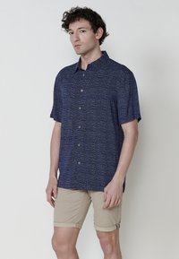 Camicia blu navy a maniche corte con un motivo testurizzato, con chiusura a bottoni e bottoni in legno naturale, abbinata a pantaloni corti beige con risvolti.